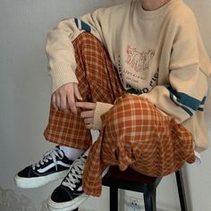 a boy sitting on a stool with his legs crossed and wearing plaid pants, a white sweater and black sneakers