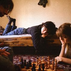 three people are playing chess on the floor in front of a bed with a wooden frame