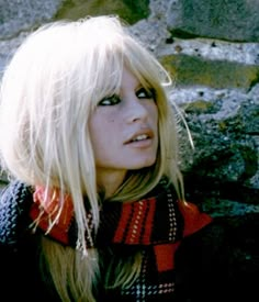 a woman with blonde hair and blue eyes wearing a scarf over her head is standing in front of some rocks