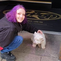 a woman kneeling down next to a small white dog with purple hair on her head