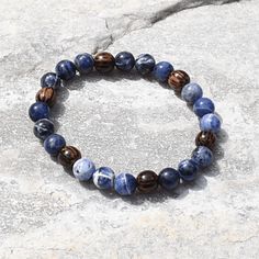 Experience the essence of harmony with the artisan-made Sodalite and Palm Wood Unisex Bracelet. This captivating accessory unites the tranquil allure of Sodalite and the rustic charm of Palm Wood, creating a wearable masterpiece that resonates with both elegance and earthy energy. Centered around the serene Sodalite beads, the bracelet showcases a palette of deep blues interwoven with delicate patterns, reminiscent of the vast expanse of a starlit sky. Sodalite is renowned for its ability to fos Spiritual Sodalite Gemstone Beaded Bracelets, Sodalite Beaded Bracelets With Natural Stones For Gift, Handmade Sodalite Round Bead Bracelets, Handmade Lapis Lazuli Bracelets For Meditation, Earthy Blue Bracelets As Gift, Earthy Blue Bracelets For Gifts, Starlit Sky, Palm Wood, Unisex Bracelets