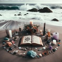 an open book sitting on top of a sandy beach next to the ocean surrounded by rocks and candles