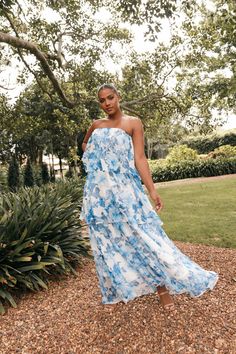 a woman wearing a blue and white dress standing in front of a bush with her hand on her hip