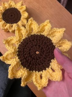 a hand holding a crocheted sunflower on top of a wooden table next to another yellow and brown flower