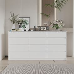 a white dresser with plants and pictures on it next to a mirror in a room