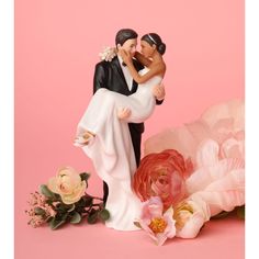 a bride and groom figurine sitting next to flowers on a pink background,