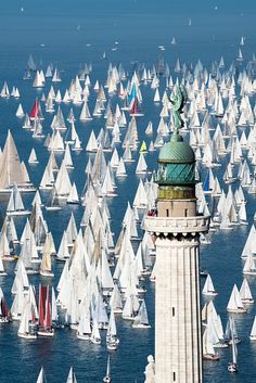 a large group of sailboats in the ocean with a lighthouse on one side and many smaller ones at the other