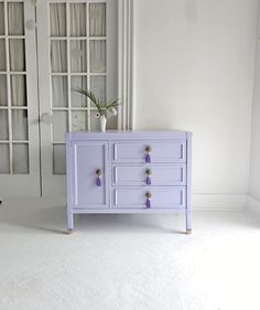 a white and purple dresser sitting in front of a window next to a vase filled with flowers