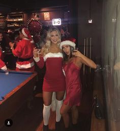 two women dressed up in santa costumes posing for the camera with pool tables and people behind them