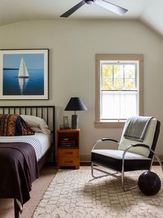 a bedroom with a bed, chair, and window in the corner that has a sailboat painting on the wall