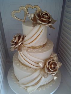 a three tiered wedding cake with gold roses on top and two hearts on the side