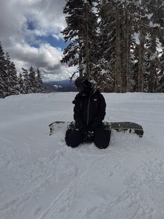 a snowboarder sitting in the snow with his board on their feet and trees behind him