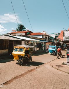 people riding motorcycles down the street in front of small shops and parked cars on the side of the road