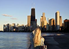 a woman sitting on the edge of a pier looking at the water and skyscrapers