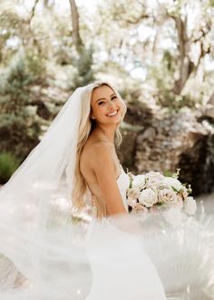 a woman in a wedding dress holding a bouquet and smiling at the camera with trees behind her