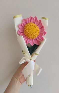 a hand holding a pink and yellow flower on top of a white paper bouquet with ribbon