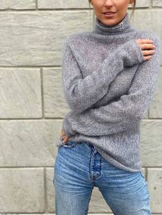 a woman wearing jeans and a turtle neck sweater poses in front of a brick wall