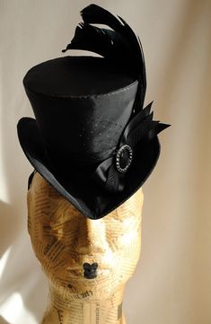 This gothic victorian mini top hat is an excellent choice for any dark cabaret event. It is covered with taffeta and embellished with various black trimmings. Two curled black rooster feathers, ribbons, a silver and black rhinestone round buckle and a just a hint of black veil. Choose between total black(pictured),blue or burgundy with black decoration. Measurements : height 4 inches( 10 cm ) front to back 9 inches ( 22 cm ) side to side 7.5 inches ( 19 cm ) This mini top hat is fully lined with Steampunk Costume Hats For Halloween Masquerade, Steampunk Fitted Brimmed Top Hat, Elegant Fitted Top Hat For Halloween, Steampunk Top Hat With Curved Brim For Costume, High Crown Top Hat For Halloween, Steampunk Headpieces For Masquerade Halloween, Steampunk Headpieces For Halloween Masquerade, Black Top Hat For Costume, Steampunk Headpiece For Halloween Costume