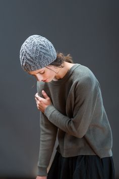 a woman standing in front of a gray wall looking at her cell phone while wearing a knitted hat