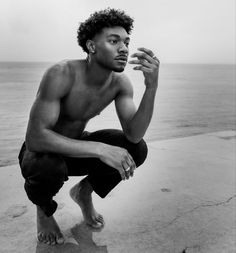 a shirtless man squatting on the edge of a cliff by the ocean with his hand in his mouth