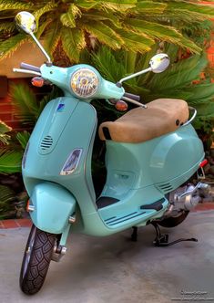 a blue scooter is parked in front of some trees and bushes, with a tan seat