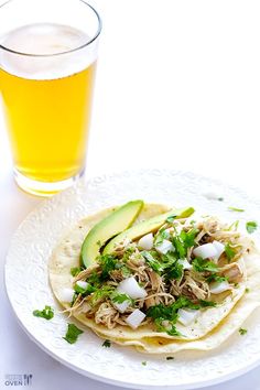 two tacos on a white plate next to a glass of beer and an avocado