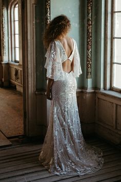 a woman standing in front of a window wearing a wedding dress with open back and sheer sleeves