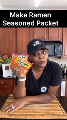 a woman sitting at a kitchen counter holding up a bag of ramen seasoned packets