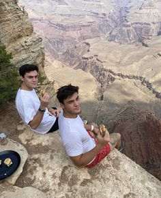 two men sitting on the edge of a cliff eating food