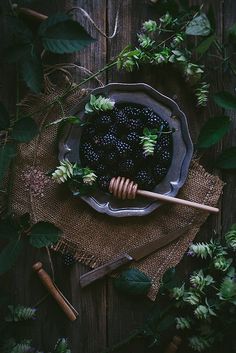 a bowl filled with blackberries next to green leaves and a wooden spoon on top of a table