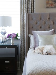 a white dog laying on top of a bed next to a night stand and lamp