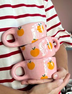 three pink cups with oranges painted on them are held in front of a striped shirt