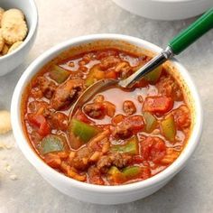 a white bowl filled with chili and meat soup next to two bowls of crackers