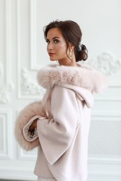 a woman wearing a pink coat with fur collar and cuffs on her shoulders, standing in front of a white wall