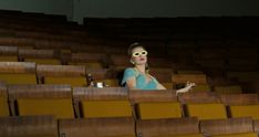 a woman wearing glasses sitting in an empty theater seat with her hand on her hip