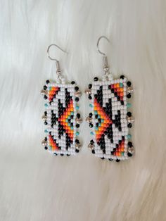 two square beaded earrings with black, white, and orange designs on them sitting on a fur surface