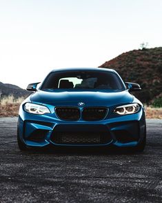 the front end of a blue car parked on top of a parking lot with mountains in the background