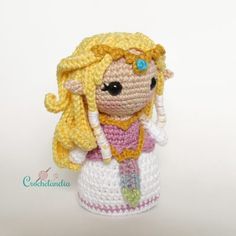 a crocheted doll with blonde hair wearing a pink dress and yellow hat is posed on a white surface