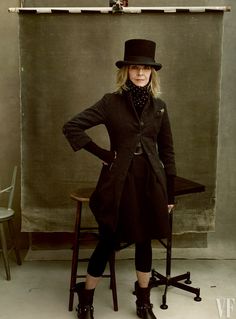 a woman in a black coat and hat standing next to a chair with her hands on her hips