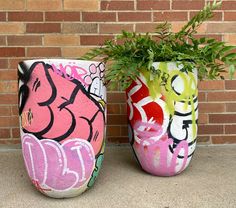 two vases with plants in them sitting on the ground next to a brick wall