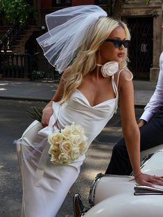 a woman in a wedding dress leaning on the hood of a white car with a veil over her head