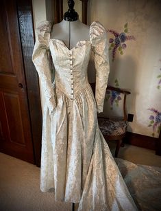 This renaissance-style dress is truly unique. The fitted princess line bodice has a square neckline front and back with a small vee at center front. This is accentuated by a mock opening in the front with "pearl" buttons, each encircled with rhinestones. Some of the buttons are missing a rhinestone. Puff sleeves, with elastic at the shoulders for wearing ease, taper to points at the wrist with a four button and loop closure. The drop waist come to a point in the front and back. Skirt is gathered. Zipper closure. What makes this truly renaissance looking is the fabric. It has gold metallic slightly raised flowers on a cream background. Dress has been cleaned and preserved since the 1991s and is in excellent condition.  Not only would this make a wonderful wedding dress, it would also be ama Princess Line, Back Skirt, Etsy Wedding Dress, Cream Background, Pearl Buttons, Drop Waist, Square Neckline, Style Dress, Puff Sleeves