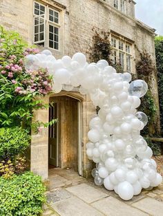 an arch made out of white balloons in front of a stone building with pink flowers