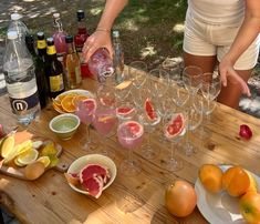 a woman is pouring wine into glasses on a wooden table with fruit and bottles in the background