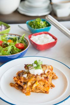 a white plate topped with lasagna covered in cheese and sauce next to bowls of salad