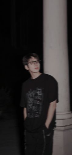 a young man wearing glasses standing next to a white pillar in the dark with his hands on his hips