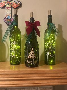 three wine bottles decorated with christmas lights and bows are sitting on a mantle in front of a cross