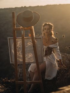 a woman is sitting next to an easel with a hat on her head and painting