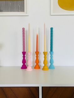 four candles are lined up on a white table with two pictures above them and one candle is brightly colored