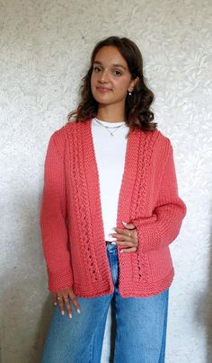 a woman standing in front of a white wall wearing a pink cardigan sweater and jeans